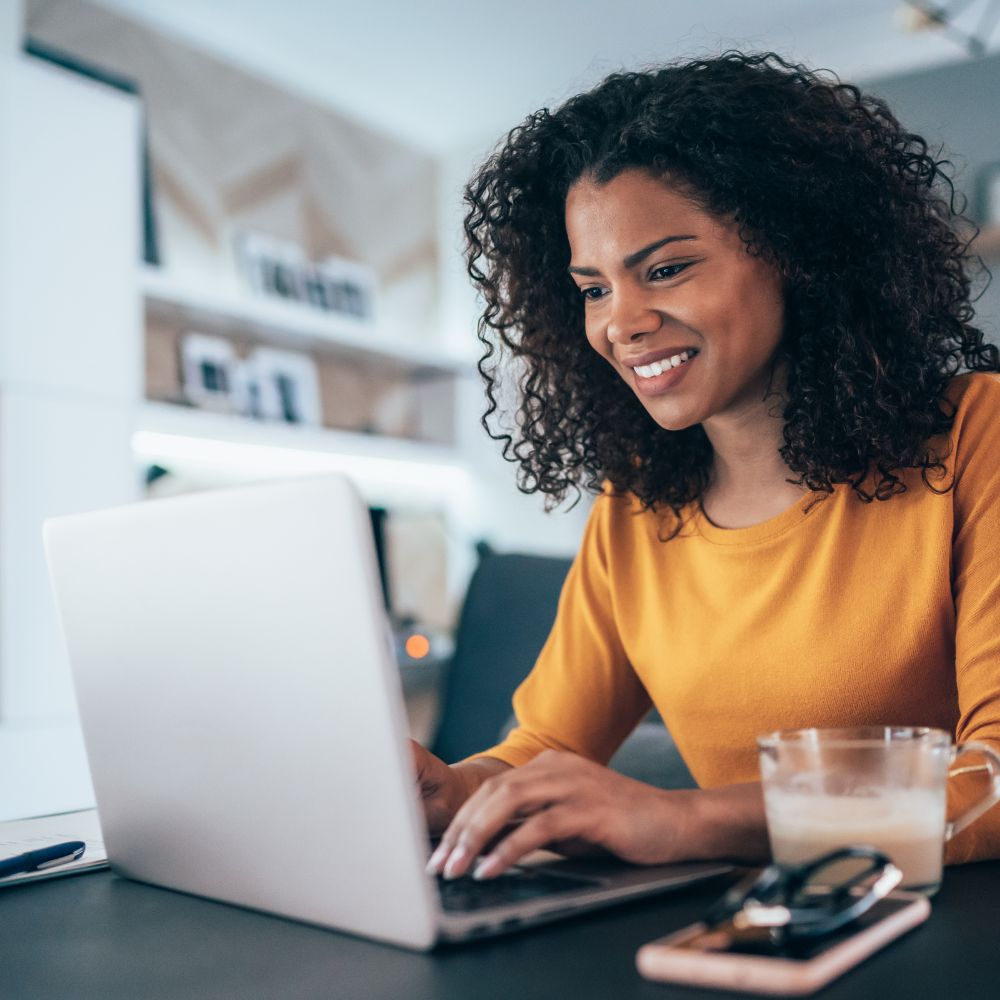 A woman with curly hair smiles while working on her laptop, showcasing a positive and engaged expression.